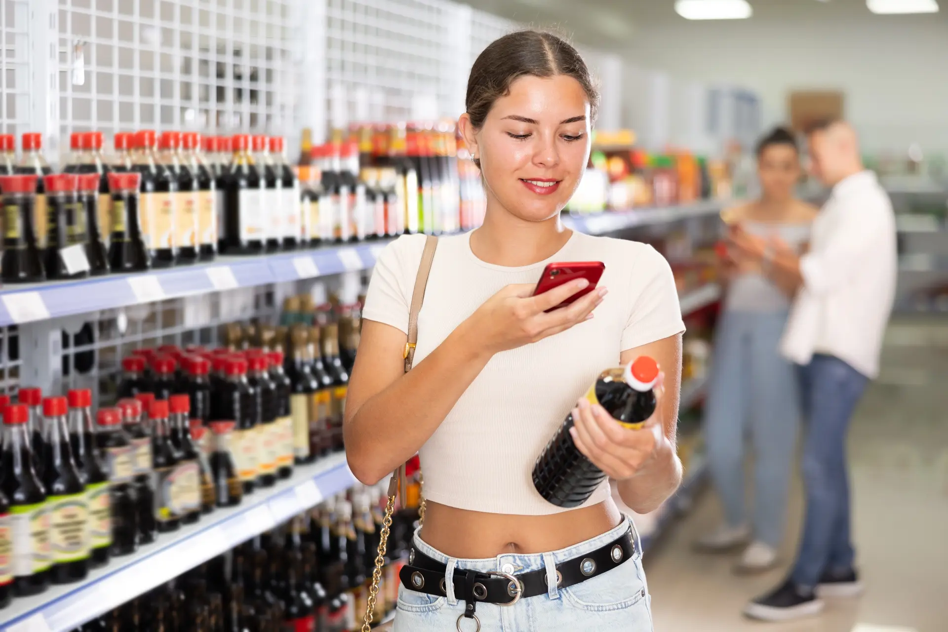 食品売り場で醤油のボトルを手に取り、スマートフォンで確認する女性